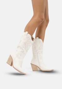 Botas de cowboy blancas con costuras decorativas, punta afilada y tacón cuadrado. Las características incluyen cremalleras laterales y una textura de cuero suave.