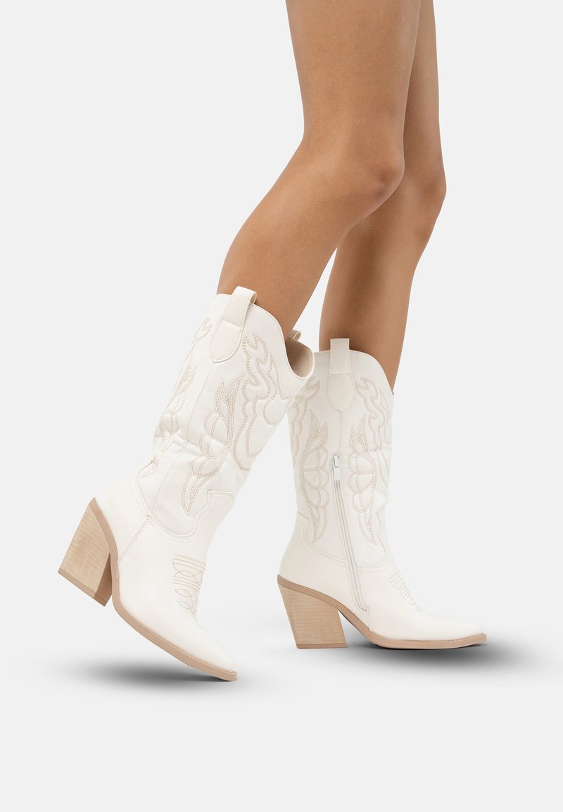 Botas de cowboy blancas con costuras decorativas, punta afilada y tacón cuadrado. Las características incluyen cremalleras laterales y una textura de cuero suave.