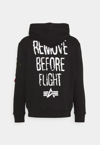 Sudadera con capucha negra que presenta un gran texto blanco "RETIRAR ANTES DEL VUELO" en la parte posterior, con un diseño de logo y un bolsillo con cremallera lateral.