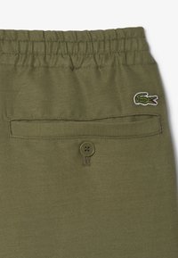 Olijfgroene broek met een elastische tailleband, een achterzak met een knop en een geborduurd krokodillenlogo. Geweven stofdetails zichtbaar.