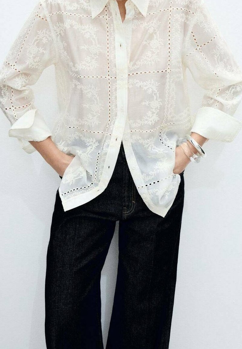 Personne portant un chemisier blanc transparent brodé avec des manches retroussées et un pantalon large noir, mains dans les poches, bracelet argenté au poignet.