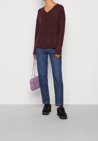 Maroon v-ringad tröja med långa ärmar, i kombination med blå raka jeans. Ytterligare detaljer inkluderar en liten lila vadderad handväska och svarta sneakers.