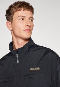 Chaqueta ligera negra con cuello alto, cierre de cremallera y dos bolsillos frontales. Presenta un parche con logo y una superficie de tela texturizada.