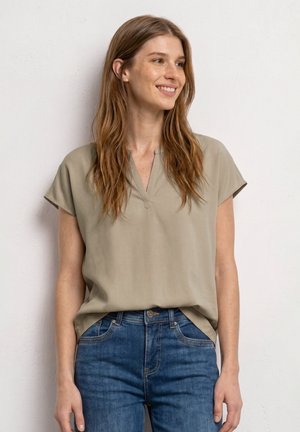 Femme souriante aux longs cheveux châtain clair portant un chemisier beige à manches courtes rentré dans un jean bleu, debout contre un mur blanc.