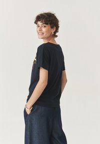 Tamsiai mėlynos spalvos trumpomis rankovėmis marškinėliai su apvalia iškirpte ir grafiniu dizainu. Modelis vilki plačias džinsines kelnes, rankas kišenėse, demonstruodamas laisvą siluetą.