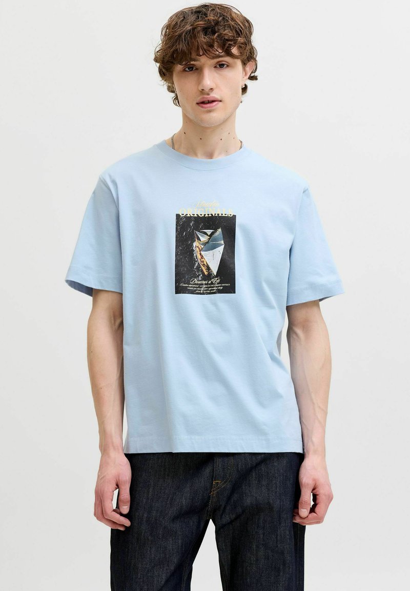 Jeune homme portant un t-shirt bleu clair avec un motif imprimé de voilier vintage et un jean foncé sur un fond uni.