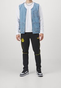 Man gekleed in zwarte Puma Borussia Dortmund broek, een witte lange mouwen shirt, een blauwe spijkerjack, zwart-witte sneakers en een zilveren ketting. 