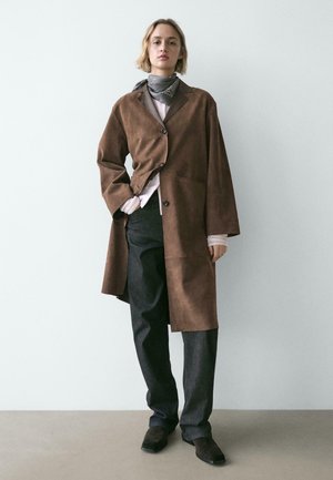 Person stående iført en brun oversized frakke, mørke bukser, gråt mønstret halstørklæde og mørke sko mod en enkel lys væg.