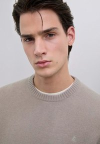 Beżowy sweter dziergany z okrągłym dekoltem w prążki, noszony na białej koszuli. Materiał wydaje się miękki, z delikatną teksturą i minimalnymi detalami.