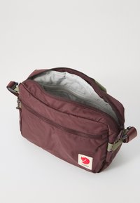 Borsa a spalla di tessuto color mogano con zip superiore, tracolla verde, tasca esterna e una patch con logo che presenta una volpe rossa sul fronte.