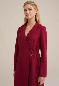 Robe rouge à manches longues, avec revers en pointe et ceinture latérale avec boucle couleur or. Texture lisse, coupe ajustée et décolleté en V profond.