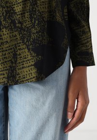 Top nero a maniche lunghe con motivo astratto verde, caratterizzato da un'orlo dritto e un tessuto morbido. Abbinato a jeans in denim azzurri.