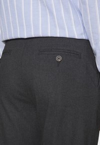 Szare, dopasowane spodnie z delikatną fakturą, wyposażone w jedną tylną kieszeń i zapięcie na guzik. Pas częściowo zakryty jest przez jasnobrązową koszulę w paski.