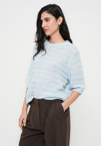 Ανοιχτό γαλάζιο πλεκτό πουλόβερ με άνετη εφαρμογή, κοντά μανίκια και υφασμένο σχέδιο. Φορεμένο με καφέ παντελόνια ψηλόμεσα.