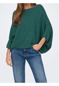 Grünes, übergroßes Stricktop mit rechteckiger Form, gerippter Struktur, weiten Ärmeln und lockerem Sitz, kombiniert mit blauen Denim-Jeans.