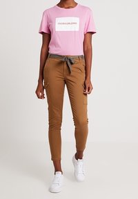 Rosa bomullströja med vit logotyp, kombinerad med bruna cargobyxor med sidofickor och ett grått midjeband, kompletterad med vita sneakers.