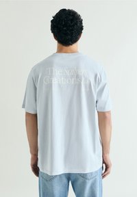 Hombre con cabello rizado y oscuro que lleva una camiseta azul clara con el texto "The Scalpers Creations.Co" en la parte trasera y vaqueros azul claro, de espaldas.