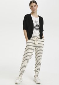 Čierny šálový kardigán, biele tričko s čiernym grafickým dizajnom a krémové pruhované tepláky so šnúrkou a strapcami. Biele tenisky.