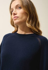 Pull bleu marine en matériau doux avec un col rond, présentant une légère transparence au niveau des coutures des épaules. Texture légère et lisse.