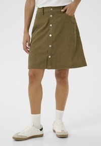 Bruine corduroy rok met verticale ribbels, voorzien van knopen aan de voorkant, zijzakken en een lichte A-lijn vorm. Gepaard met witte sneakers.