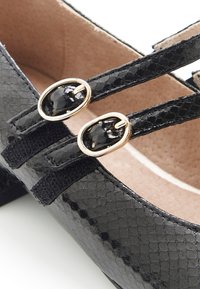 Zapatos de charol negros con una superficie texturizada, dos correas negras y acentos ovalados dorados cerca de la parte superior del zapato. El interior es beige.