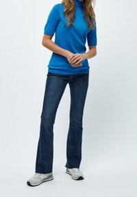 Blå kortärmad polotröja, tillsammans med mörkblå utsvängda jeans och ljusgrå sportskor. Bakgrunden är helt vit.