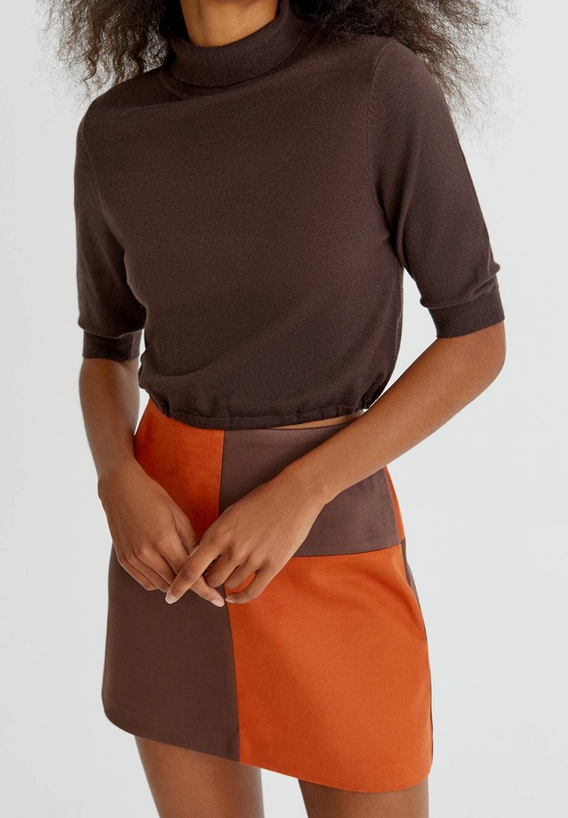 Femme portant un haut marron à manches courtes à col roulé et une jupe taille haute en blocs de couleur orange et marron, debout les mains jointes.