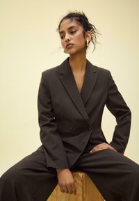 Blazer marrone a righe sottili a doppio petto con pantaloni coordinati. Tessuto liscio, vestibilità su misura, rever a punta e due bottoni anteriori.