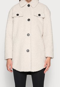 Cappotto beige peloso con colletto, due tasche frontali e bottoni neri. Tessuto con texture e vestibilità ampia.