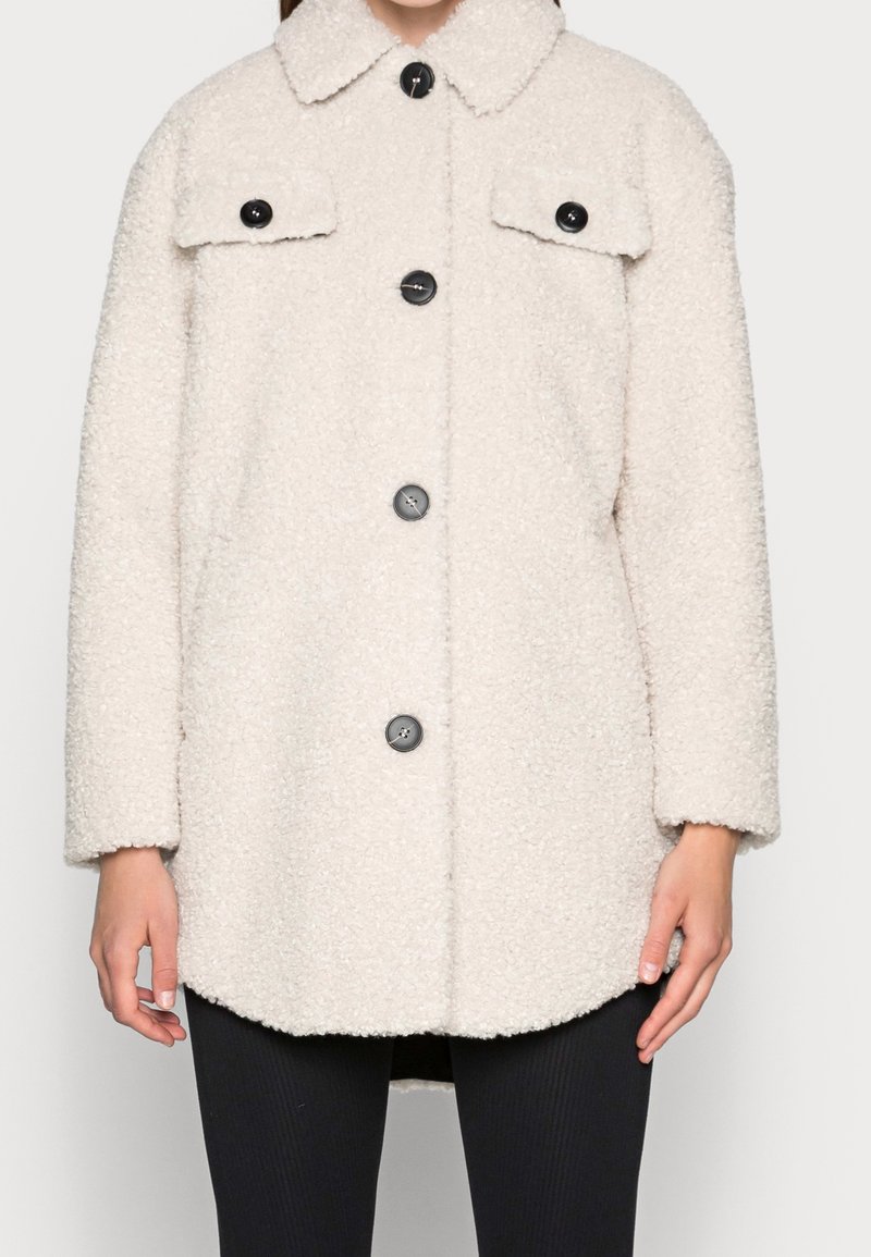Cappotto beige peloso con colletto, due tasche frontali e bottoni neri. Tessuto con texture e vestibilità ampia.
