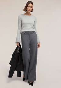 Grå texturerad tröja med långa klockärmar, kombinerad med högmidjade, vida mörkgrå byxor. Svart läderjacka hålls i ena handen.