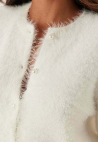 Primo piano di un cardigan bianco soffice con bottoni di perla indossato da una persona, che mostra la trama e il colletto.