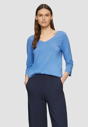 Hemelsblauw langemouwen top met een V-hals ontwerp, gemaakt van zachte stof; gedragen met marineblauwe wijde broek, handen in de zakken.