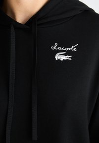 Černá mikina s kapucí a stahovací šňůrkou, která na hrudi obsahuje bílé výšivky loga "Lacoste" a designu krokodýla. Hladký povrch.