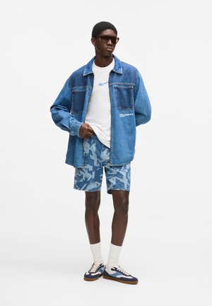 Fiatal férfi sötét napszemüvegben, túlméretezett kék farmerdzsekiben, fehér pólóban, mintás farmer rövidnadrágban, fehér zokniban és kék tornacipőben, fehér háttér előtt.