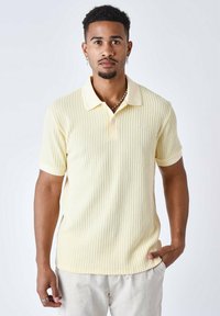 Camisa polo amarilla con textura suave, con cuello y mangas cortas, combinada con pantalones de color claro. Detalle sutil del logo.