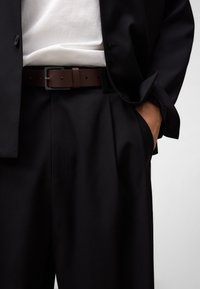 Svart kavaj över en vit skjorta, med ett brunt läderbälte med rektangulär metallspänne, och skräddarsydda svarta byxor med fickor.