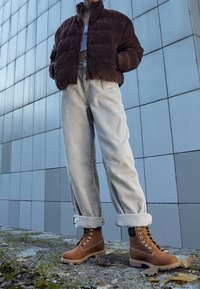 Veste matelassée en velours côtelé marron, haut bleu clair, pantalon en velours côtelé beige remonté aux chevilles, et bottines marron robustes avec des lacets dorés.