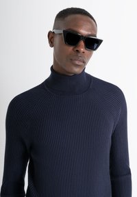 Maglione a collo alto ribbed blu navy con design aderente e maniche lunghe, abbinato a occhiali da sole neri angolari con lenti scure.