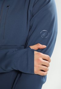 Mørkeblå langærmet jakke lavet af blødt, glat materiale; har et logo på den venstre ærme og en lynlåslomme på brystet.