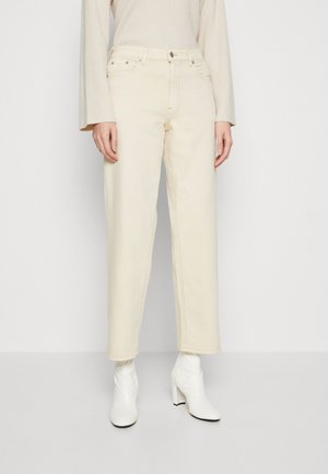 Osoba ubrana w kremowe spodnie typu straight-leg, kremowy prążkowany sweter oraz białe botki na obcasie, stojąca na tle jednolitego jasnego tła.