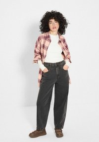 Różowa koszula flanelowa w kratę założona na białą koszulkę z długim rękawem, w zestawie z luźnymi ciemnymi dżinsami i brązowymi butami na sznurowadła. Teksturowane loki dodają objętości.