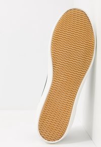 Semelle de chaussure avec un motif texturé ondulé de couleur gomme, bordée d'un bord en caoutchouc blanc, posée sur un fond clair.