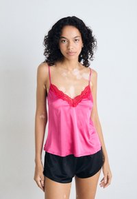 Camisole en satin rose avec des bordures en dentelle rouge, dotée de fines bretelles et d'une coupe décontractée. Associée à un short noir. Texture lisse et design léger.