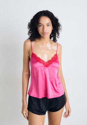 Camisole en satin rose avec des bordures en dentelle rouge, dotée de fines bretelles et d'une coupe décontractée. Associée à un short noir. Texture lisse et design léger.