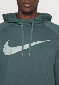 Hoodie vert foncé avec une poche kangourou à l'avant, des cordons de serrage et un grand logo Nike argenté. Présente une texture côtelée et des manches raglan.