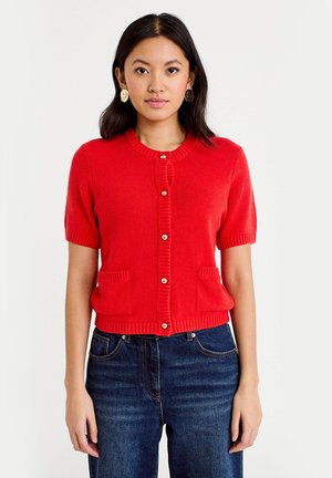 Donna che indossa un cardigan rosso brillante a maniche corte con bottoni e due tasche frontali, abbinato a jeans blu scuro su uno sfondo uniforme.
