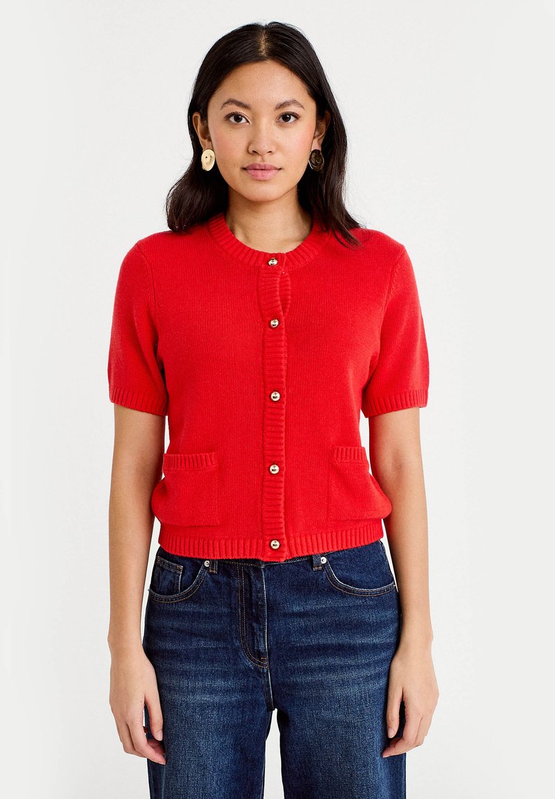 Femme portant un cardigan rouge vif à manches courtes boutonné, avec deux poches frontales, et un jean bleu foncé, posant devant un fond uni.