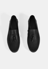Schwarze Leder Slipper mit strukturierten Oberflächen, dekorativen Quasten und aufwendigen Nähten entlang der Kanten. Minimalistisches Design.