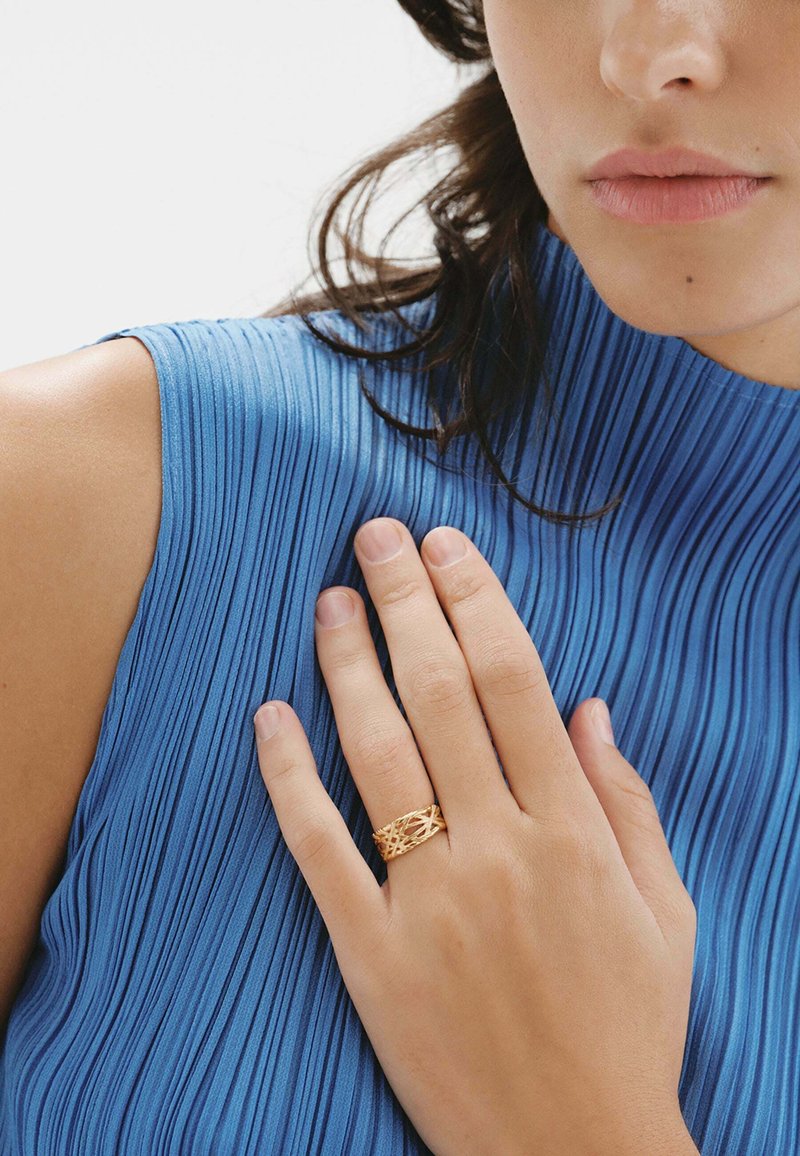 Anillo de oro con un intrincado diseño trenzado. La mano está posicionada contra una tela azul plisada de alto cuello y textura.